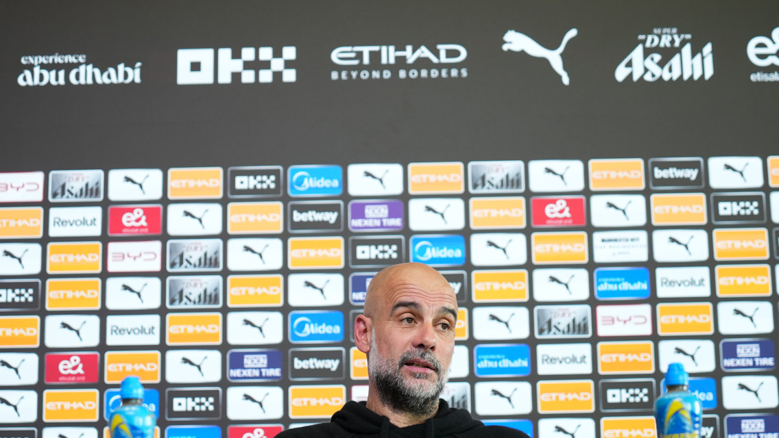 A person sitting at a press conference with a backdrop featuring various brand logos.