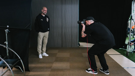 A photographer with a tattoo on their arm takes a picture of a person standing against a vertical striped backdrop. The person is wearing a dark jacket with a Puma logo, beige pants, and white shoes.