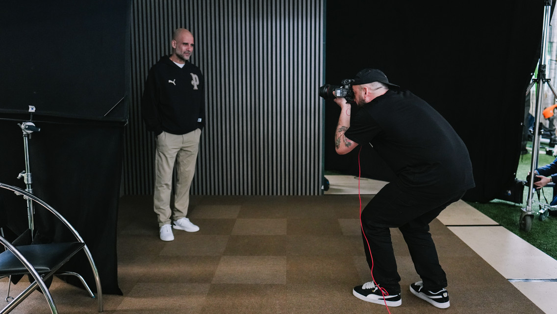 A photographer with a tattoo on their arm takes a picture of a person standing against a vertical striped backdrop. The person is wearing a dark jacket with a Puma logo, beige pants, and white shoes.