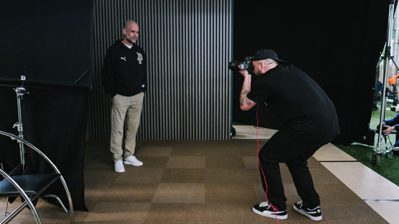 A photographer with a tattoo on their arm takes a picture of a person standing against a vertical striped backdrop. The person is wearing a dark jacket with a Puma logo, beige pants, and white shoes.