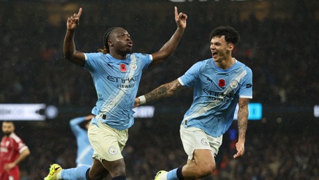 Two Manchester City players celebrating a goal on the field, wearing light blue jerseys with club logos.
