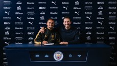 Two individuals at a Manchester City signing event in front of a sponsor backdrop.