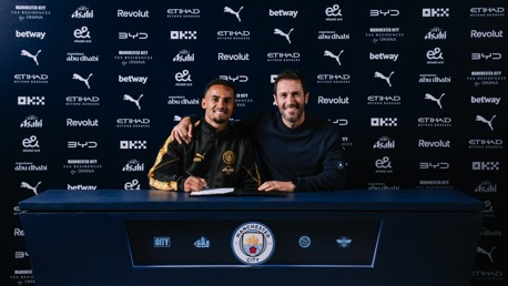 Two individuals at a Manchester City signing event in front of a sponsor backdrop.