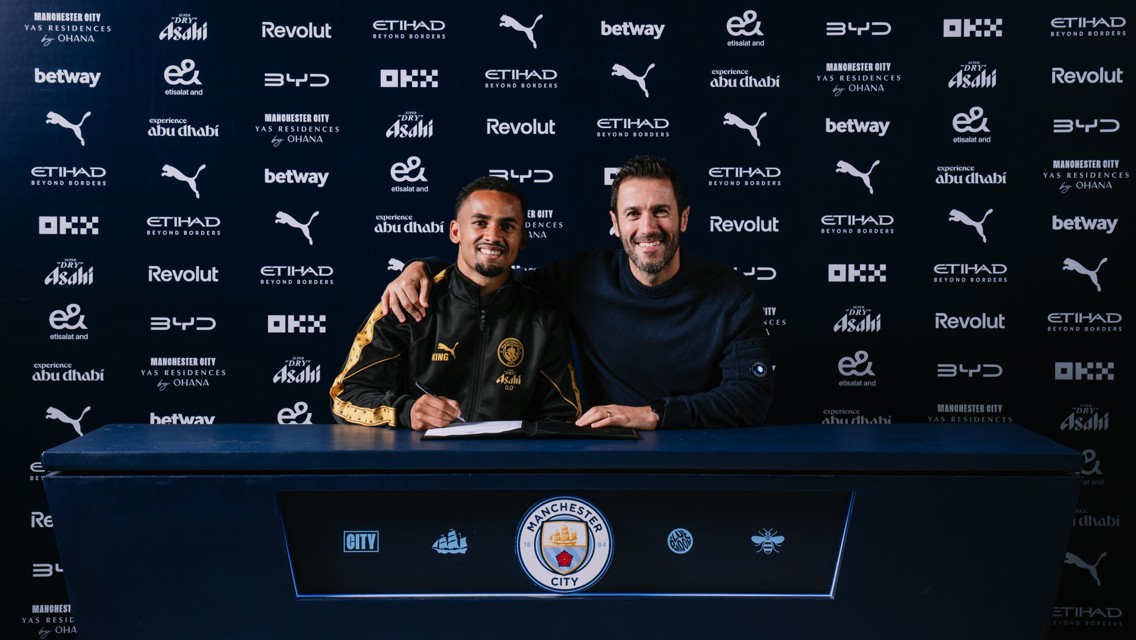 Two individuals at a Manchester City signing event in front of a sponsor backdrop.