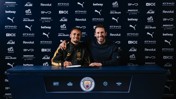 Two individuals at a Manchester City signing event in front of a sponsor backdrop.