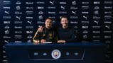 Two individuals at a Manchester City signing event in front of a sponsor backdrop.