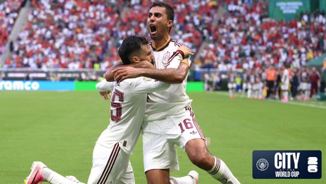 Two soccer players in white jerseys celebrate a goal on the field. One player kneels and the other stands with arms outstretched. The stadium is crowded with fans. Manchester City and World Cup logos are visible.