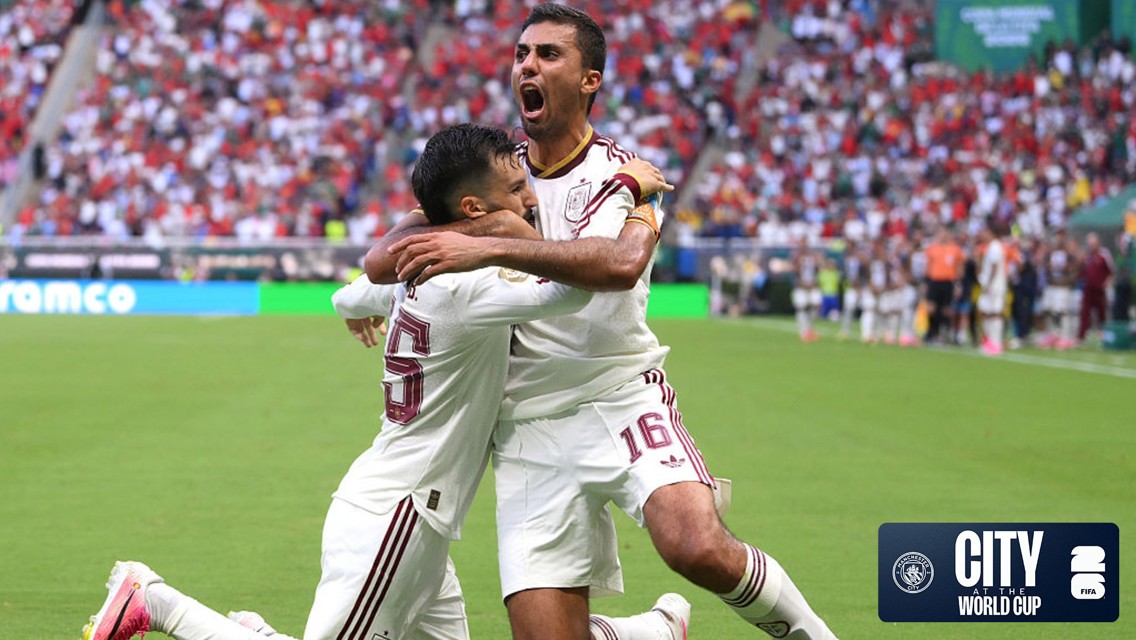 Two soccer players in white jerseys celebrate a goal on the field. One player kneels and the other stands with arms outstretched. The stadium is crowded with fans. Manchester City and World Cup logos are visible.
