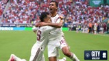 Two soccer players in white jerseys celebrate a goal on the field. One player kneels and the other stands with arms outstretched. The stadium is crowded with fans. Manchester City and World Cup logos are visible.