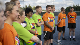 A group of people wearing green and orange shirts stand on a soccer field, showcasing pride flags on their uniforms. A soccer ball with pride colors is visible.