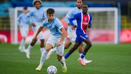 Soccer players in light blue jerseys competing against players in red and blue on a field, focusing on a young player dribbling the ball.