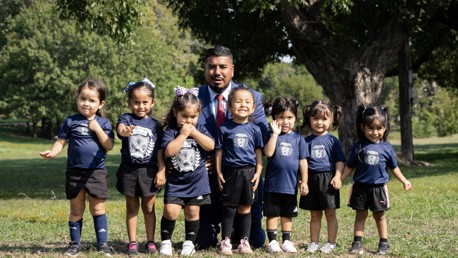 Group photo of a coach with seven young children wearing matching soccer uniforms in a park setting.