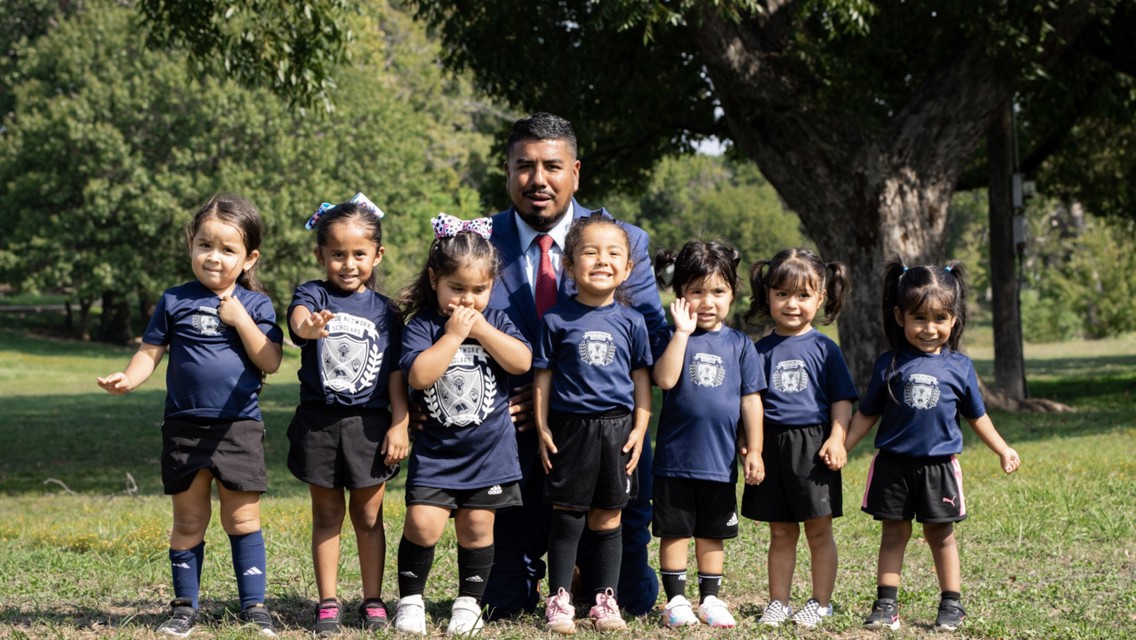 Group photo of a coach with seven young children wearing matching soccer uniforms in a park setting.