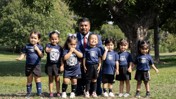 Group photo of a coach with seven young children wearing matching soccer uniforms in a park setting.