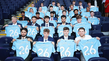 A group of young individuals in suits holding Manchester City jerseys with 'Class of 26' written on them, seated in a stadium.