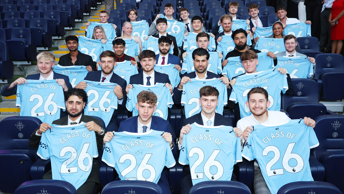 A group of young individuals in suits holding Manchester City jerseys with 'Class of 26' written on them, seated in a stadium.