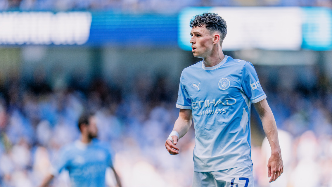 A Manchester City player wearing the team's light blue home kit on the field during a match. The player's face is blurred.