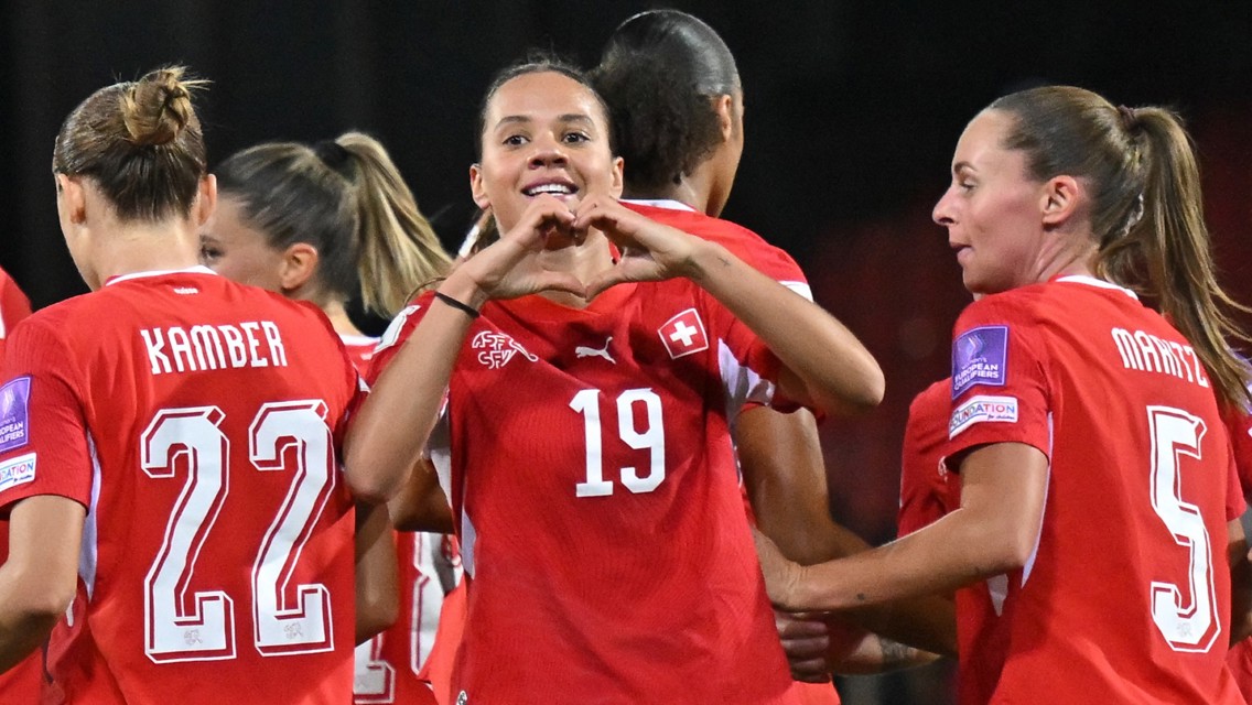 Swiss women's soccer team celebrating a goal. The player wearing jersey number 19 forms a heart shape with her hands.