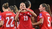 Swiss women's soccer team celebrating a goal. The player wearing jersey number 19 forms a heart shape with her hands.