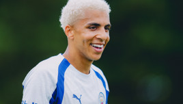A smiling Manchester City football player with bleached blond hair, wearing the team training kit.