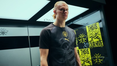 A football player wearing a Manchester City and Puma shirt in front of bee-themed posters.