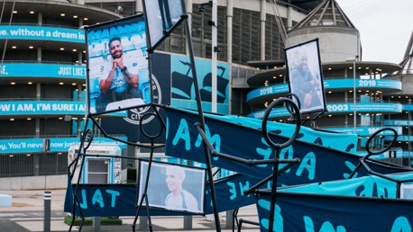 An outdoor exhibit at Etihad Stadium featuring portraits and signage related to Manchester City with stadium architecture in the background.