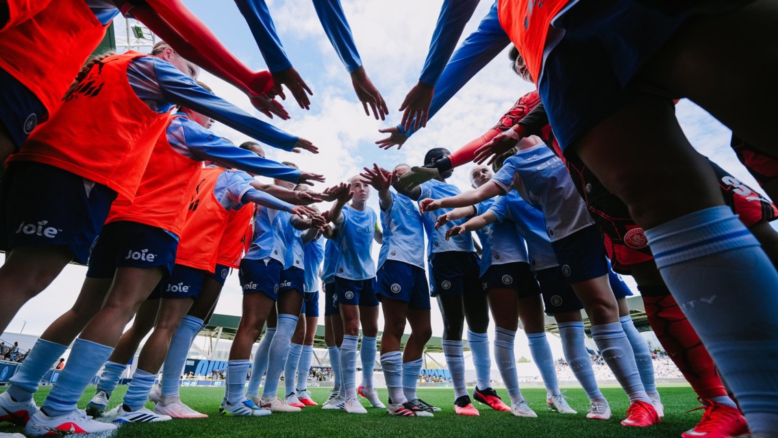 A group of soccer players wearing Manchester City kits huddling together on the field before a match, with arms extended towards the center.