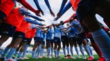 A group of soccer players wearing Manchester City kits huddling together on the field before a match, with arms extended towards the center.