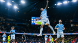 A Manchester City player jumps in celebration after scoring a goal, with teammates and a scoreboard showing 2-0 in the background.