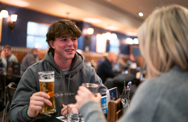 Two people at a pub, talking and holding drinks, with a blurred background of other patrons.