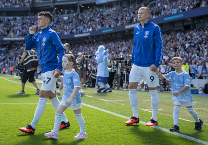 Manchester City players walking on the field at Etihad Stadium with children as mascots. The players are wearing blue tracksuits and the children are in Manchester City kit.
