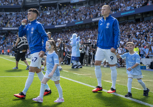 Manchester City players walking on the field at Etihad Stadium with children as mascots. The players are wearing blue tracksuits and the children are in Manchester City kit.