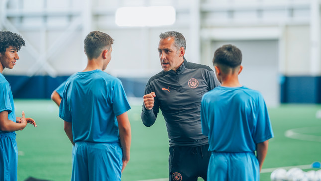 A coach is instructing young football players wearing blue kits on an indoor football field. Soccer balls are visible in the background.