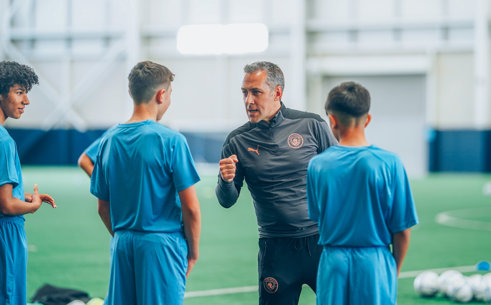 A coach is instructing young football players wearing blue kits on an indoor football field. Soccer balls are visible in the background.
