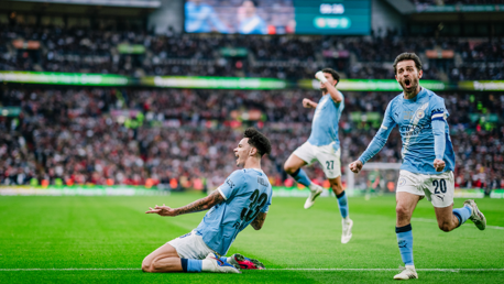 Soccer players in blue jerseys celebrating a goal on a field with large crowd in the background.