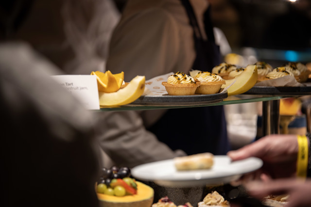 A close-up view of lemon tarts with fruit garnishes on a buffet table, with people serving themselves in the background.