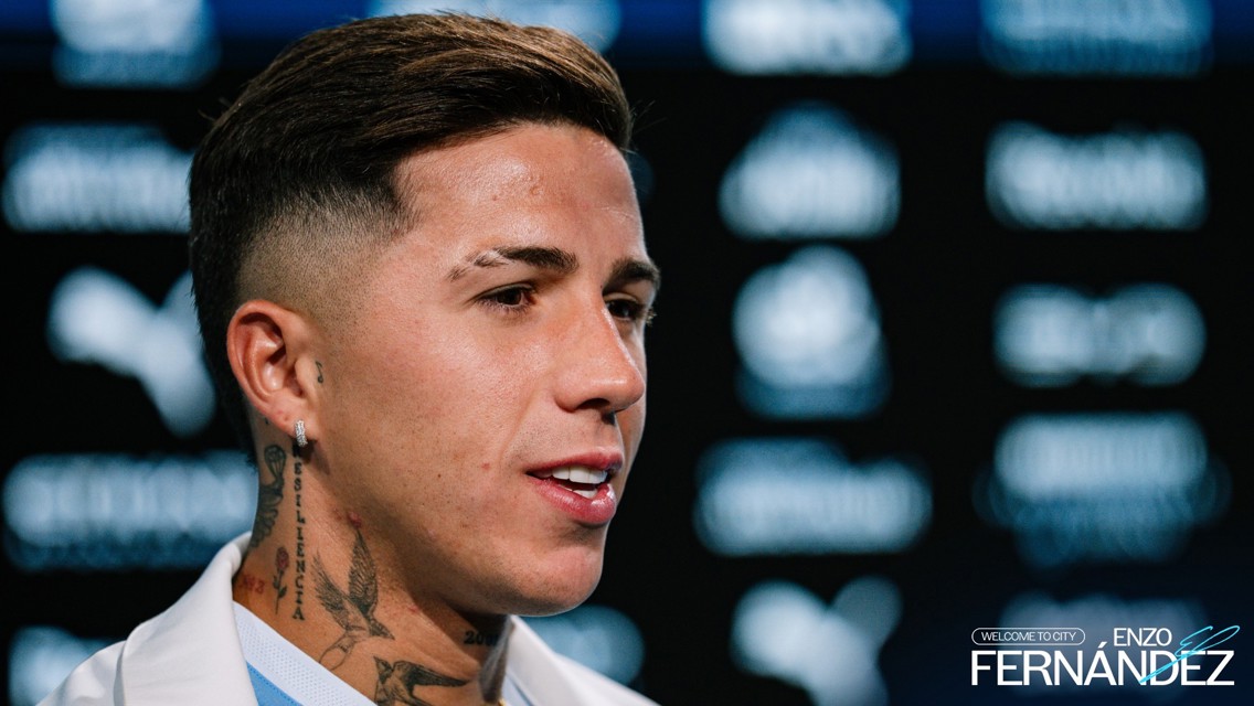A person with tattoos and earrings against a blurred backdrop of logos. The text 'Welcome to City' and 'Enzo Fernandez' are visible.