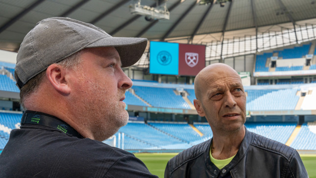 Two people with blurred faces standing in a stadium with large screens displaying logos of Manchester City and West Ham United.