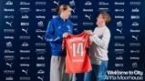 Two individuals are holding a Manchester City shirt with the name 'Moorhouse' and the number '14'. The background includes various sponsor logos. Text reads 'Welcome to City Anna Moorhouse'.