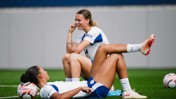 Two female footballers in Manchester City training kit sitting on a grass field, laughing and relaxing, with footballs nearby.
