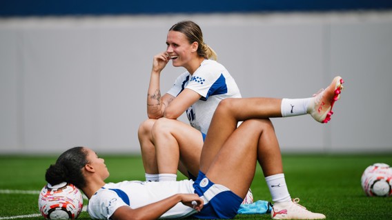 Two female footballers in Manchester City training kit sitting on a grass field, laughing and relaxing, with footballs nearby.
