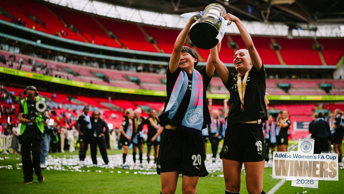 Two female football players celebrate winning Adobe Women's FA Cup 2026 at Wembley Stadium, holding the trophy aloft with medals around their necks. Confetti is scattered on the field.