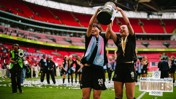 Two female football players celebrate winning Adobe Women's FA Cup 2026 at Wembley Stadium, holding the trophy aloft with medals around their necks. Confetti is scattered on the field.
