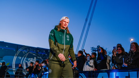 A person in a Manchester City tracksuit arrives with fans taking photos. The team bus is in the background with Manchester City logos visible.