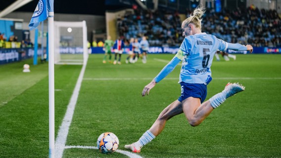 A player in a Manchester City jersey taking a corner kick during a soccer match at night. The goal area is visible in the background with other players and spectators.