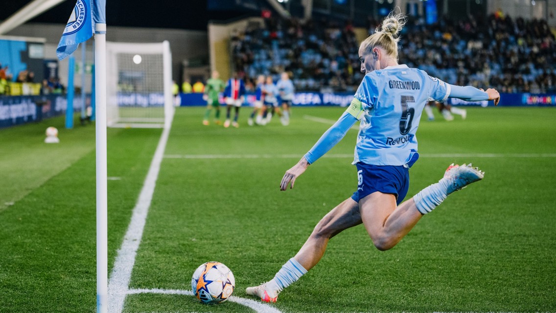 A player in a Manchester City jersey taking a corner kick during a soccer match at night. The goal area is visible in the background with other players and spectators.