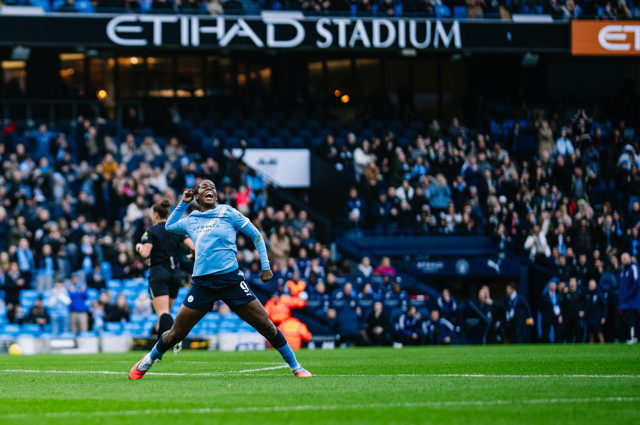 A Manchester City player celebrates during a game at Etihad Stadium. The scene captures the player in motion with fans in the background and the stadium signage visible.