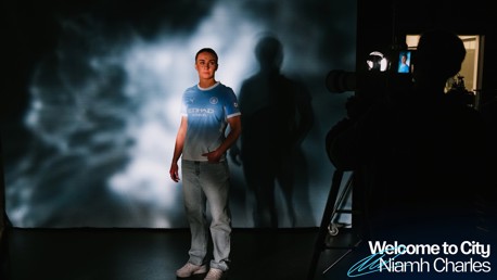 A person in a Manchester City jersey is standing in front of a camera, with a light projection creating a patterned background. The text 'Welcome to City Niamh Charles' is visible.