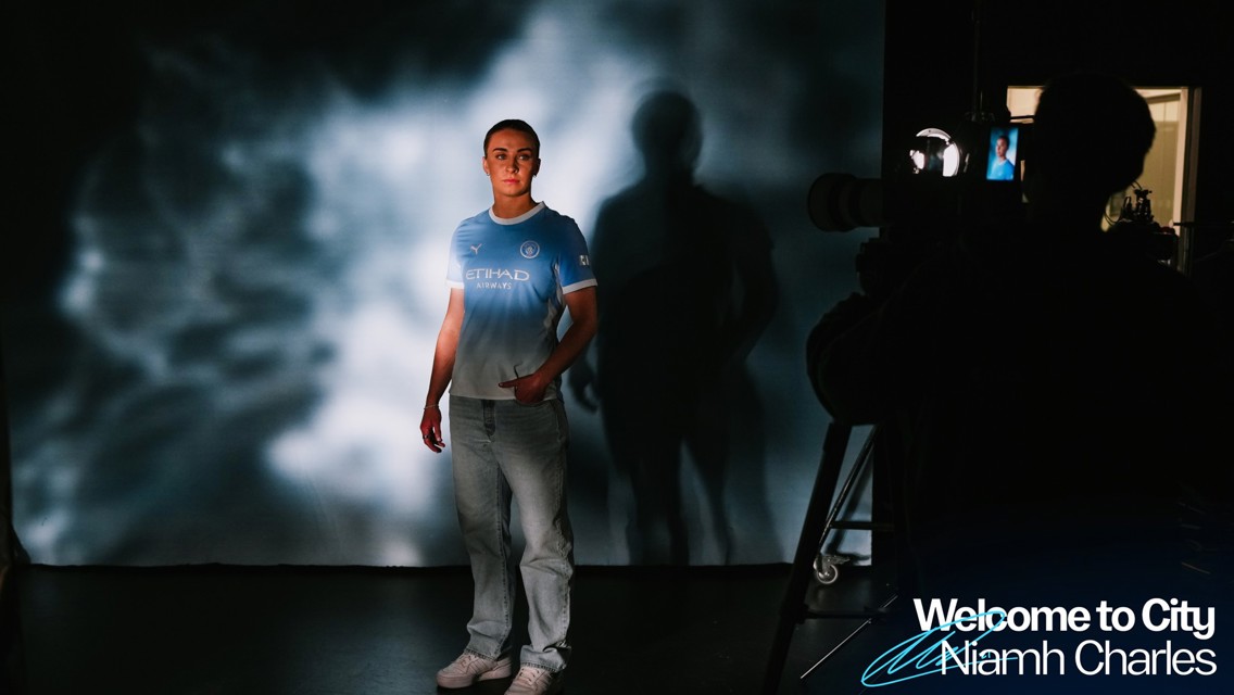 A person in a Manchester City jersey is standing in front of a camera, with a light projection creating a patterned background. The text 'Welcome to City Niamh Charles' is visible.