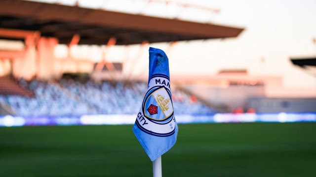 A Manchester City corner flag on a soccer field with blurred stands in the background.